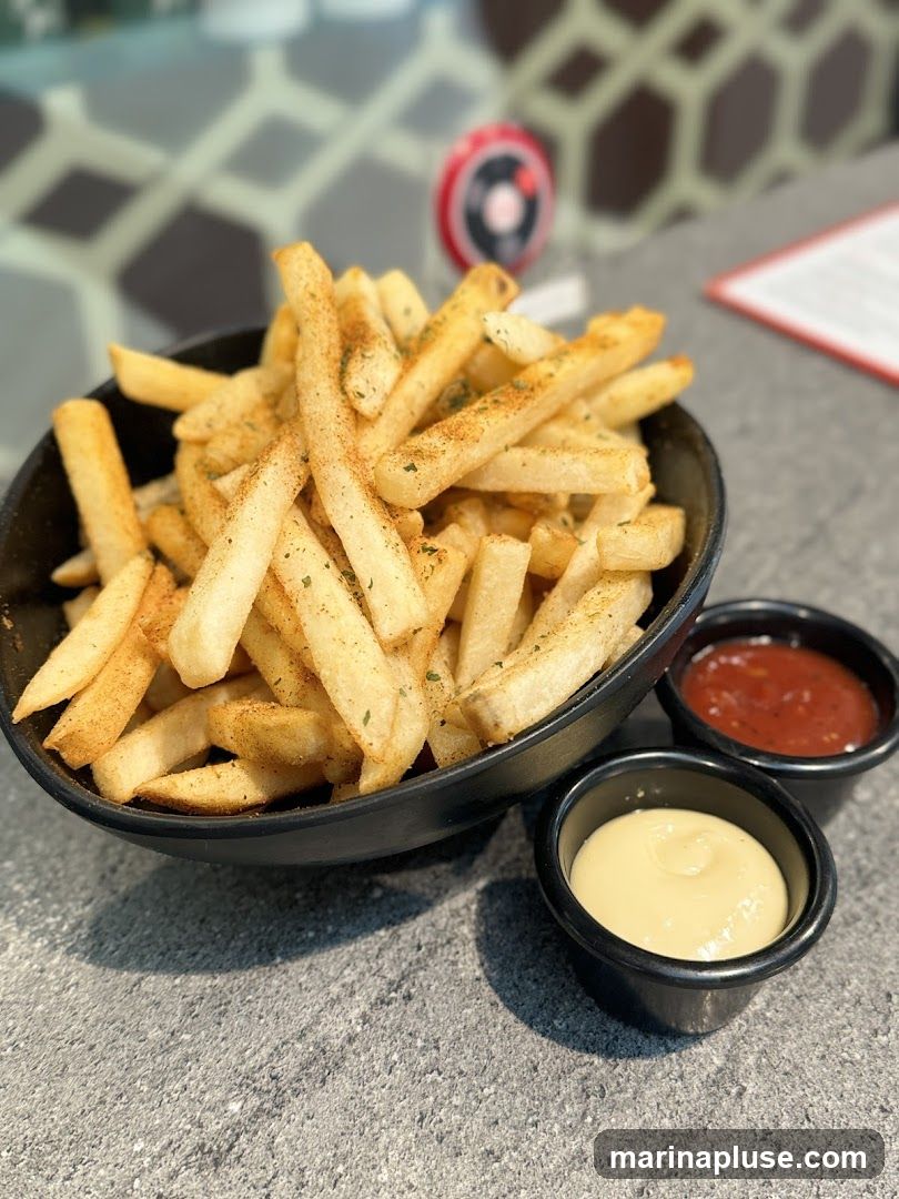 A close-up shot of a bowl of seasoned french fries, served with two small dipping bowls, one containing ketchup and the other a creamy yellow sauce, possibly mayonnaise or aioli. The fries are golden brown and sprinkled with herbs and spices. The background is blurred, showing a patterned wall and a red menu or document.
