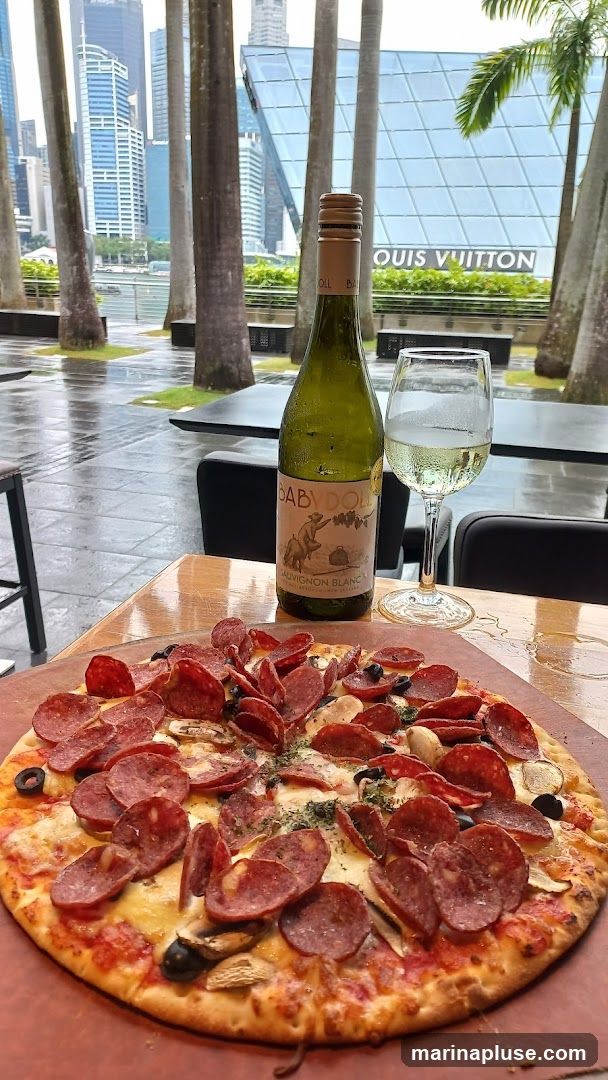 A close-up of a pepperoni pizza and a bottle of white wine with a glass of wine on a wooden table. In the background, palm trees and modern buildings are visible, including a building with a Louis Vuitton sign. The ground appears wet, suggesting it may have rained.