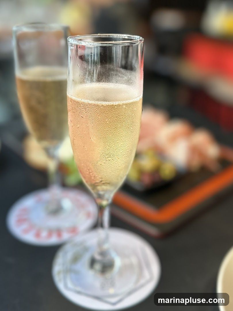 Two glasses of champagne are in focus, with condensation on the glass of the foreground drink. In the background, a charcuterie board with sliced meats and olives is visible. The setting appears to be a restaurant or bar.