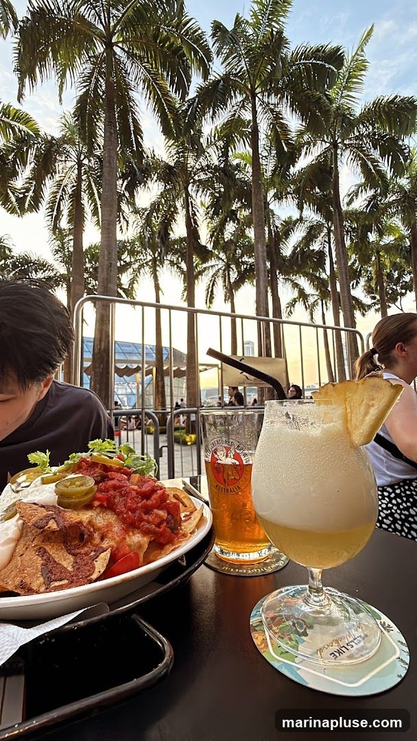 A plate of nachos topped with salsa, cheese, and jalapeños is served alongside a glass of beer and a cocktail with a pineapple wedge. The setting appears to be an outdoor seating area with numerous palm trees and a view of a modern building in the background. The lighting suggests it is late afternoon or early evening.