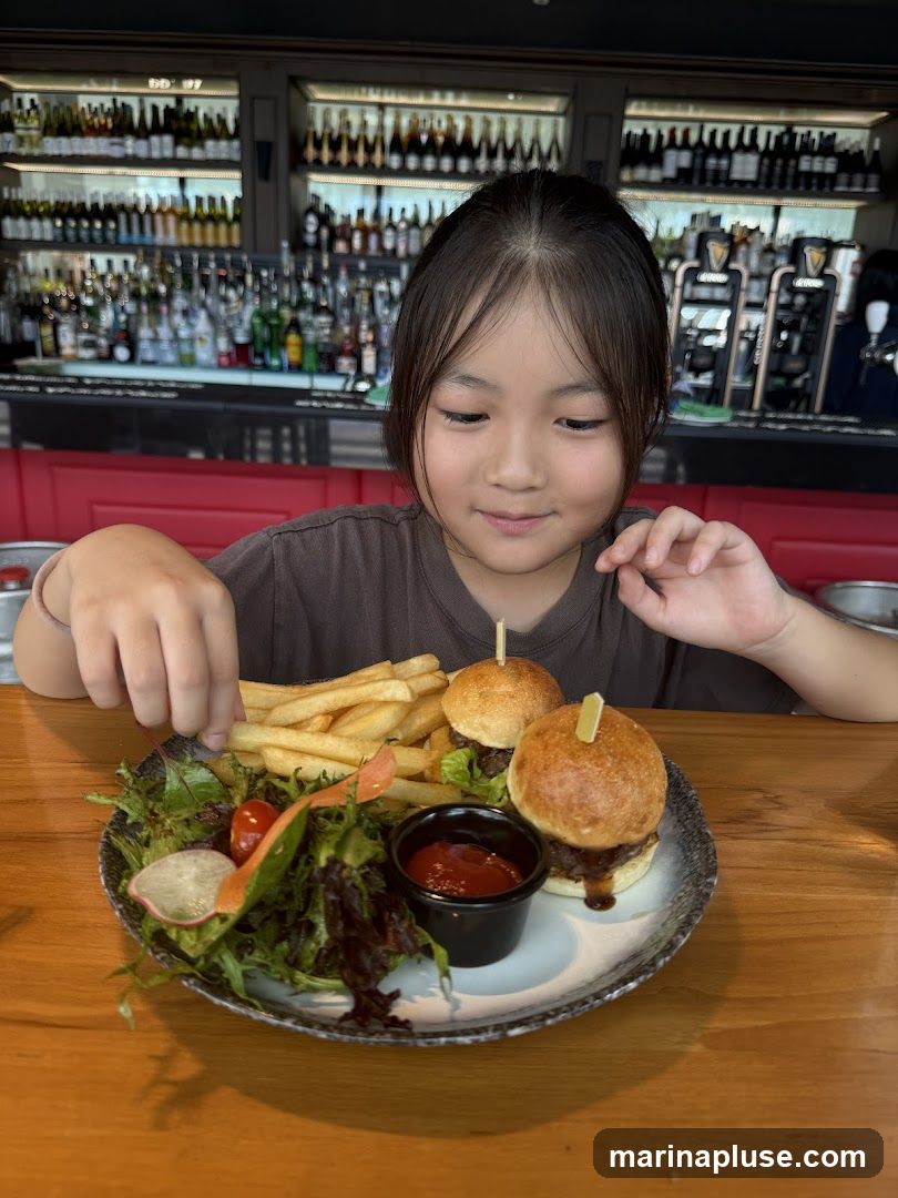 A young girl looks down at a plate of food in front of her. The plate contains two sliders, a side of french fries, a small salad with cherry tomatoes and radish, and a cup of ketchup. In the background, a bar is stocked with bottles of alcohol and has Guinness taps.