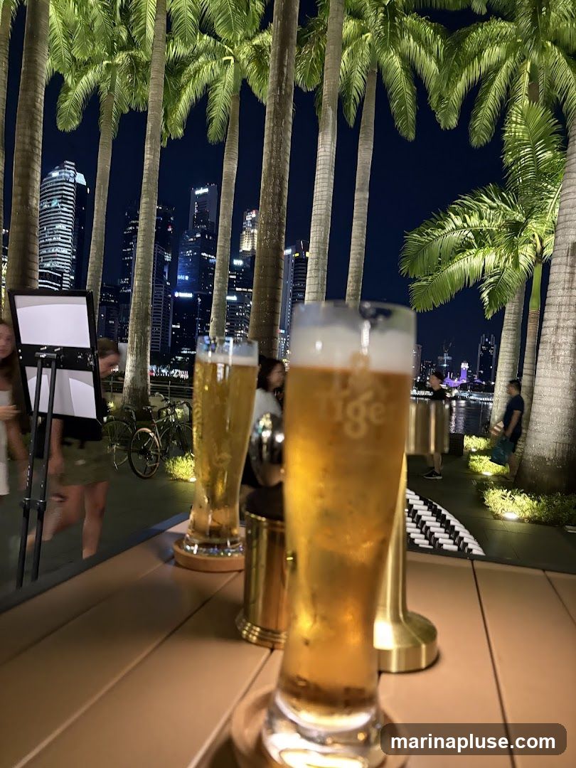 Two glasses of beer are on a table in the foreground. Behind them, palm trees line a walkway with a city skyline in the distance. People are visible in the background, and a bicycle is parked near a stand with a sign. The scene is set at night.