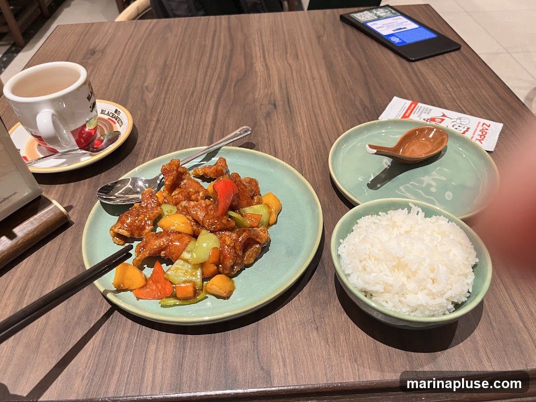 A plate of sweet and sour pork with pineapple, bell peppers, and carrots is served with a bowl of white rice, a cup of tea, and chopsticks on a wooden table. A small dish with a spoon and a packet of Zappy are also visible.