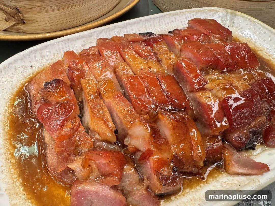 A close-up shot of sliced char siu pork, a popular Cantonese roasted pork dish, served on a white rectangular plate. The pork is glazed with a sweet and savory sauce, giving it a glossy sheen. The meat is a vibrant red and pink, with some pieces showing a slightly charred edge. In the background, out of focus, are bamboo steamers.