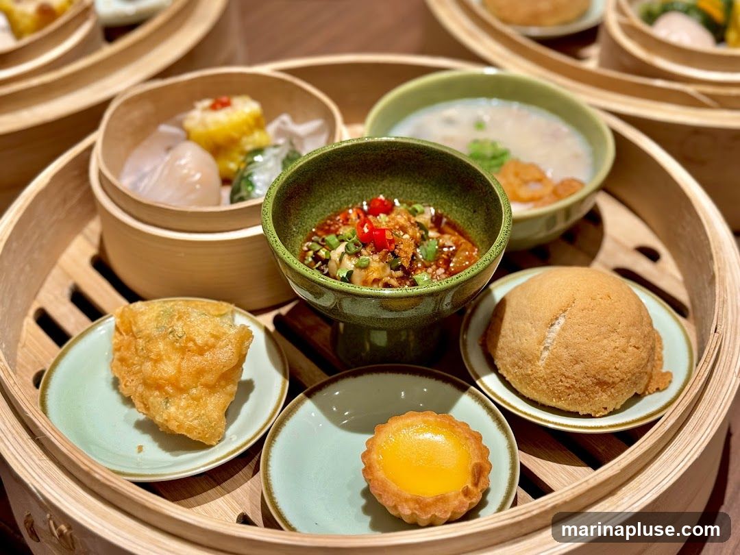 A close-up shot of a dim sum platter featuring various steamed and fried delicacies. In the center, a small green bowl holds a dipping sauce with chili and herbs. To its left, a golden-brown fried item sits on a light green plate. To the right, a fluffy bun is presented on a similar plate. Below the sauce bowl, a mini egg tart with a bright yellow filling is on its own small plate. In the background, bamboo steamers hold translucent dumplings and yellow-topped siu mai, along with a bowl of congee or soup with shrimp.