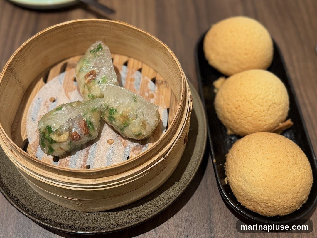 A close-up shot of a dim sum meal. In a bamboo steamer, there are three translucent dumplings filled with green vegetables and nuts. To the right, on a black plate, are three golden-brown baked buns.