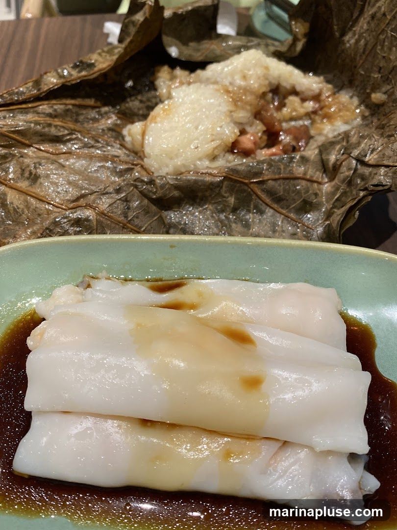 A close-up shot of two popular dim sum dishes. In the foreground, delicate white rice noodle rolls are served in a dark savory sauce. In the background, a portion of sticky rice wrapped in a lotus leaf is visible, revealing its contents of rice, meat, and possibly beans.