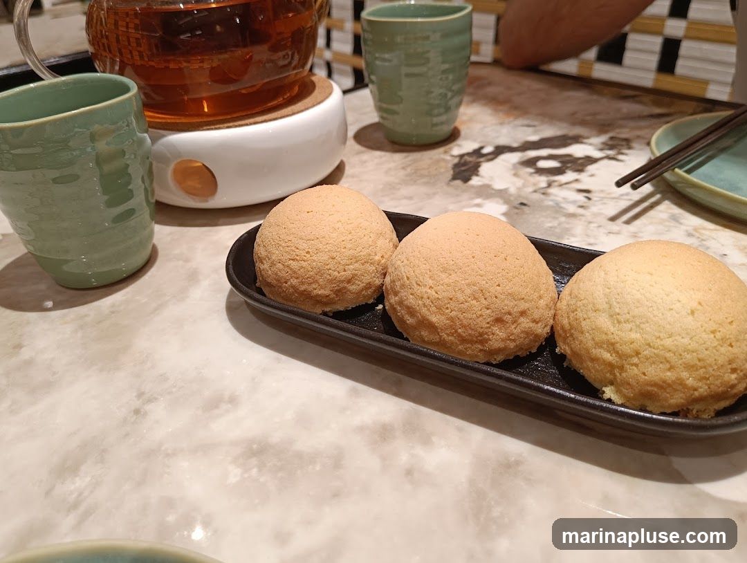 Three golden-brown baked buns are served on a black rectangular plate. To the left, a green ceramic cup and a glass teapot filled with amber tea sit on a white warmer. The table has a marble-like surface. In the background, chopsticks rest on a light blue plate, and a person's arm is visible.