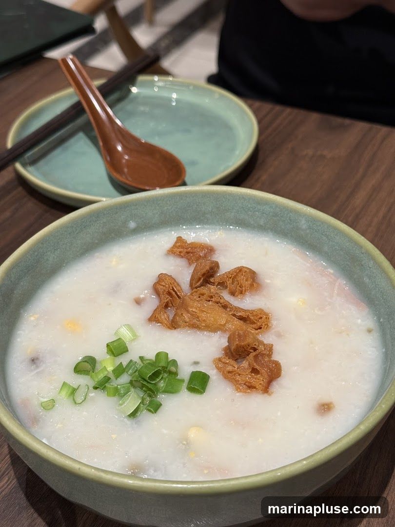 A close-up shot of a bowl of congee, a type of rice porridge, topped with fried dough fritters and chopped scallions. A spoon and chopsticks rest on a small plate in the background.