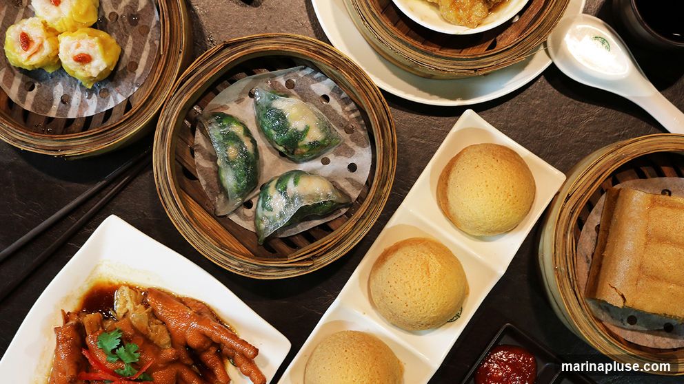 A top-down view of a variety of dim sum dishes served in bamboo steamers and on white plates. Visible items include shrimp dumplings (siu mai), steamed buns, chicken feet in sauce, and a rolled pastry. Chopsticks and a serving spoon are also present.