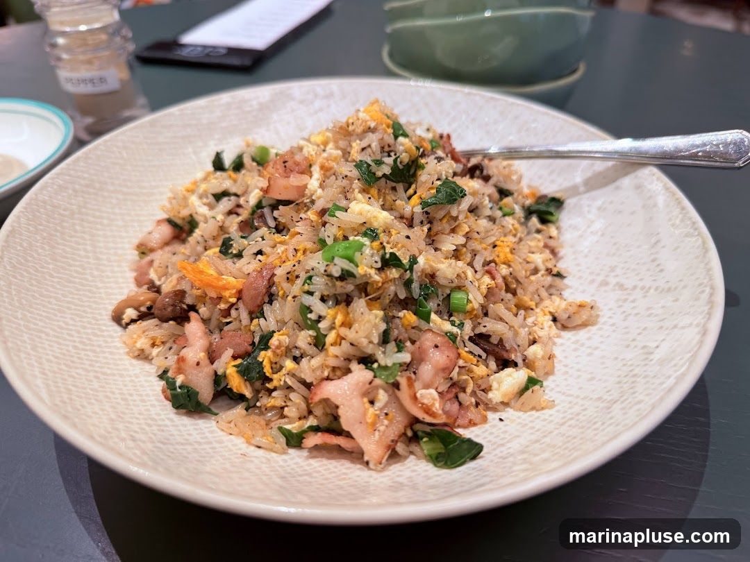 A plate of fried rice is shown, featuring rice mixed with scrambled eggs, chopped green onions, pieces of shrimp, and bacon. A fork rests on the edge of the plate. In the background, a salt shaker labeled 'PEPPER' and a stack of green bowls are visible.