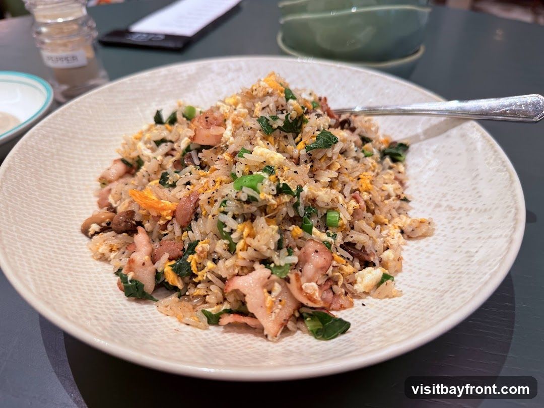 A plate of fried rice is shown, featuring rice mixed with scrambled eggs, chopped green onions, pieces of shrimp, and bacon. A fork rests on the edge of the plate. In the background, a salt shaker labeled 'PEPPER' and a stack of green bowls are visible.