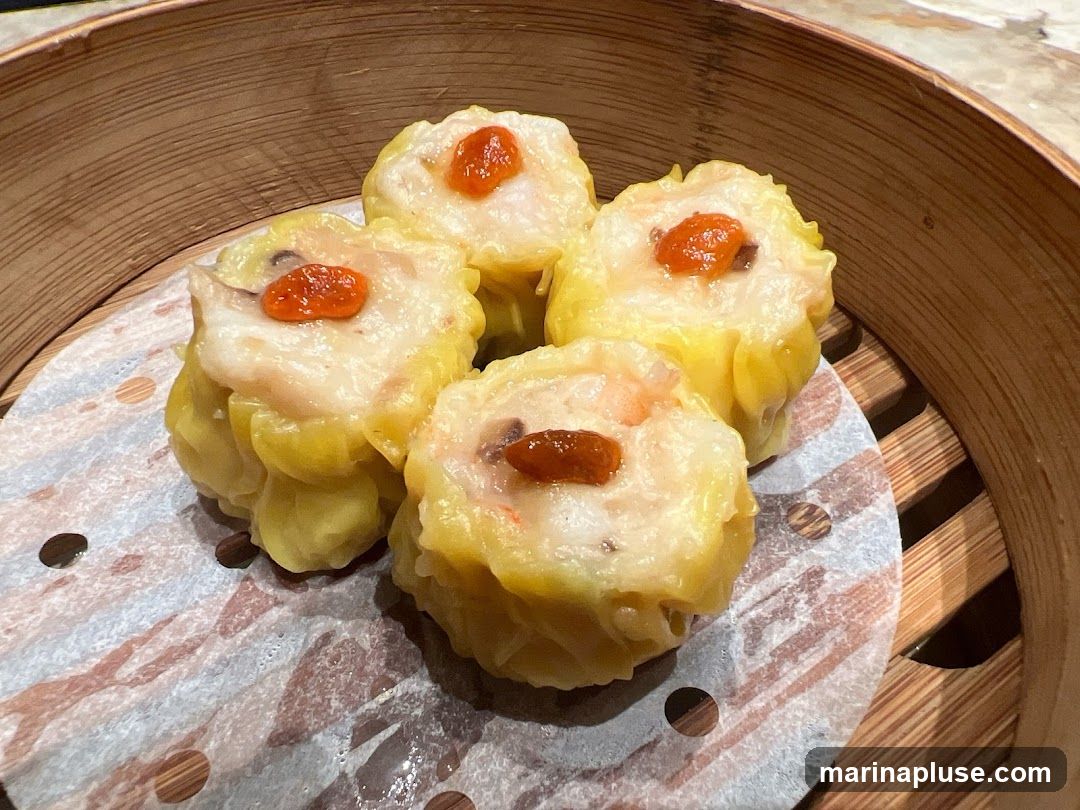 A close-up shot of four siu mai dumplings in a bamboo steamer. The dumplings are topped with a small piece of red goji berry or similar garnish. The steamer is lined with patterned parchment paper.