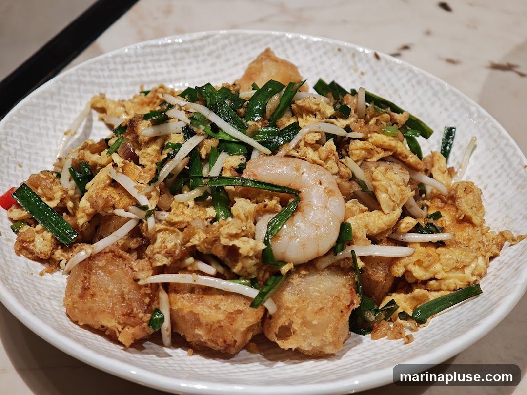 A close-up shot of a white, textured plate filled with a stir-fried dish. The dish contains scrambled eggs, chopped green onions, bean sprouts, and several pieces of fried taro or radish cake. A single large shrimp is prominently placed on top. The food appears to be seasoned and lightly browned.