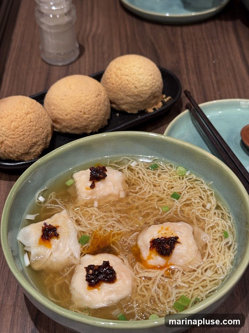A close-up shot of a bowl of wonton noodle soup with four large wontons topped with chili oil and scallions. The soup contains thin egg noodles and is garnished with chopped green onions. To the left, on a black plate, are three golden-brown baked buns with a textured, crispy crust. A salt shaker is visible in the background.