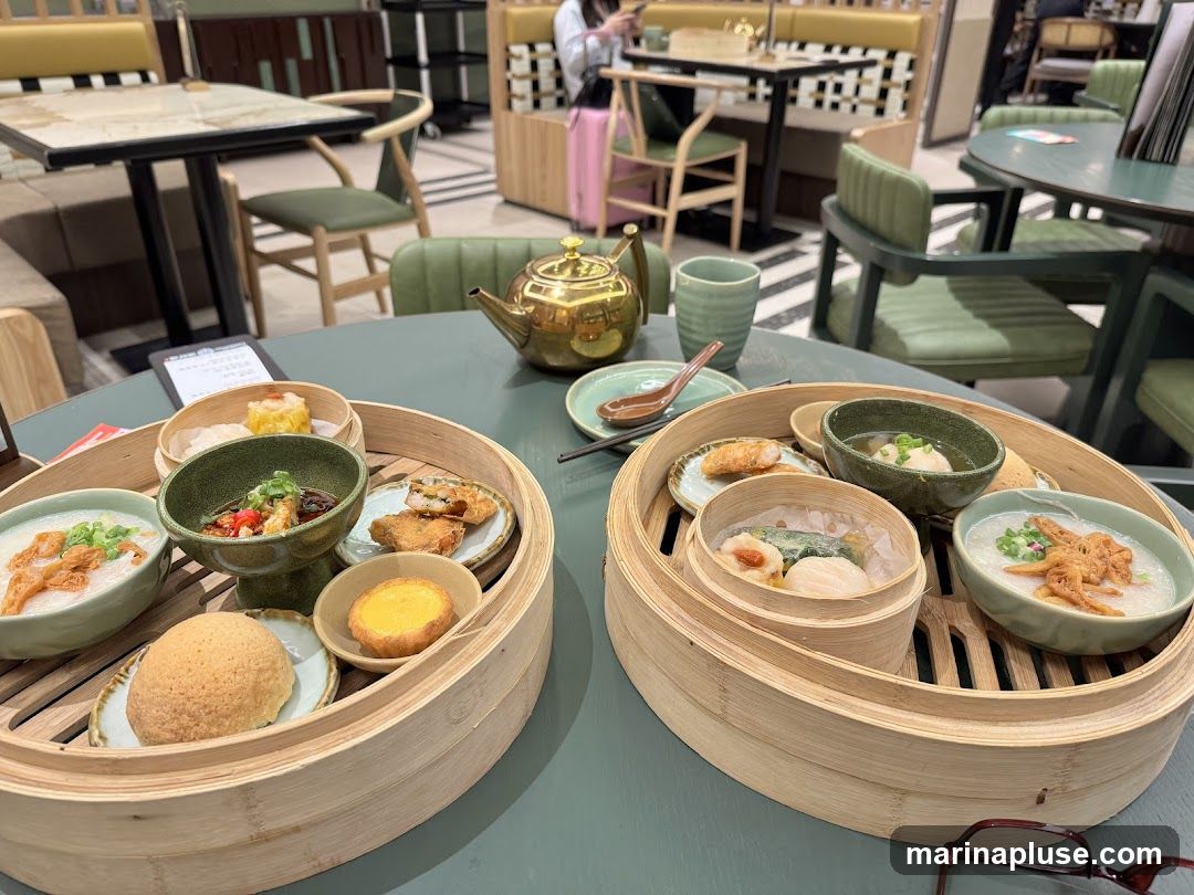 A close-up view of two bamboo steamers filled with various dim sum dishes, including congee, dumplings, fried items, and a tart. A golden teapot and a green teacup are visible in the background, along with dining tables and chairs in a restaurant setting.