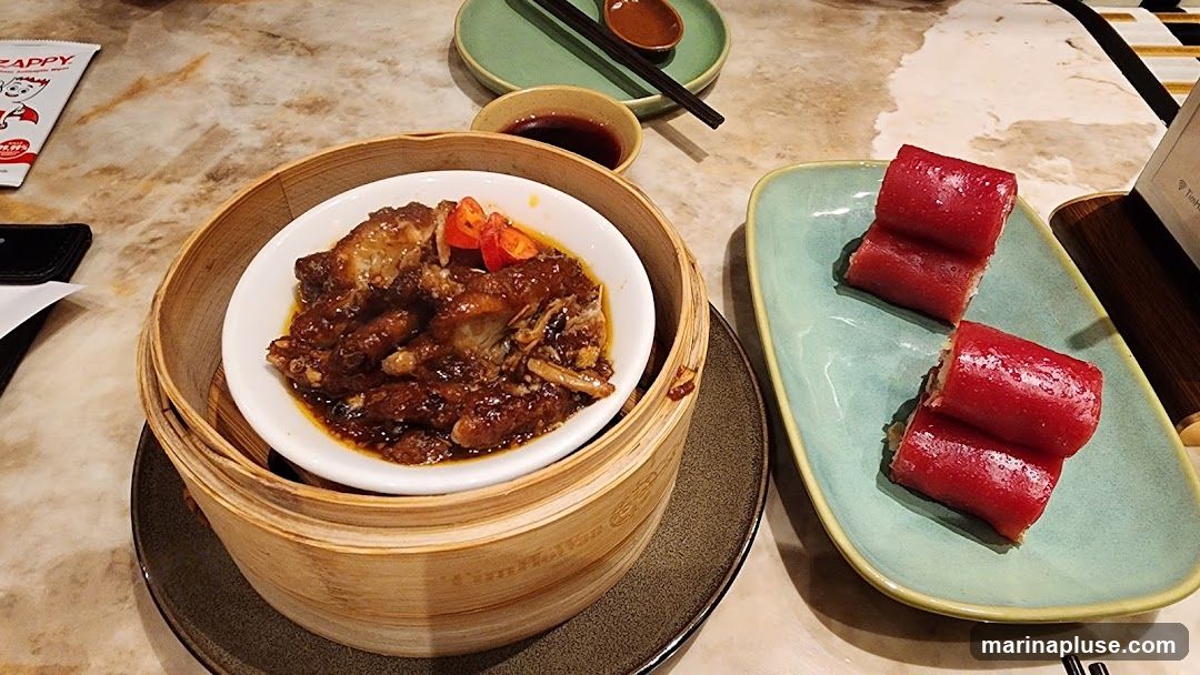 A close-up shot of a dim sum meal. In a bamboo steamer, there are braised chicken feet in a savory sauce, garnished with red chili and scallions. To the right, a light green plate holds several pieces of red rice noodle rolls. A small dish of soy sauce and chopsticks are visible in the background. A packet of antiseptic wipes with a cartoon character is on the left.