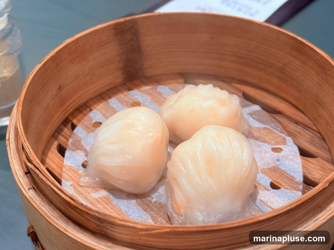 Three steamed shrimp dumplings (har gow) are presented in a bamboo steamer basket lined with patterned paper. The dumplings are translucent with visible shrimp filling. A blurred menu or document is visible in the background.