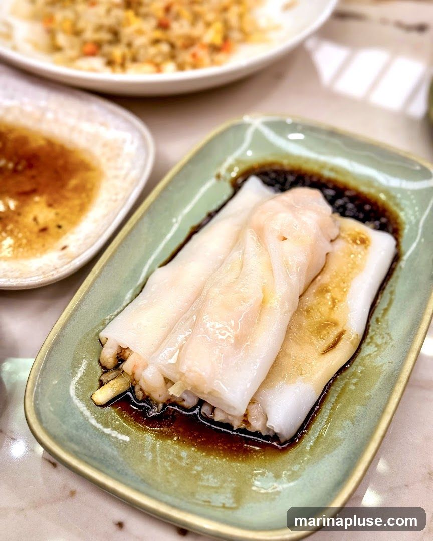 A close-up shot of three shrimp rice noodle rolls served in a light green rectangular dish. The rolls are translucent and filled with shrimp, resting in a dark soy sauce. In the background, a plate of fried rice and a small dish of dipping sauce are visible.