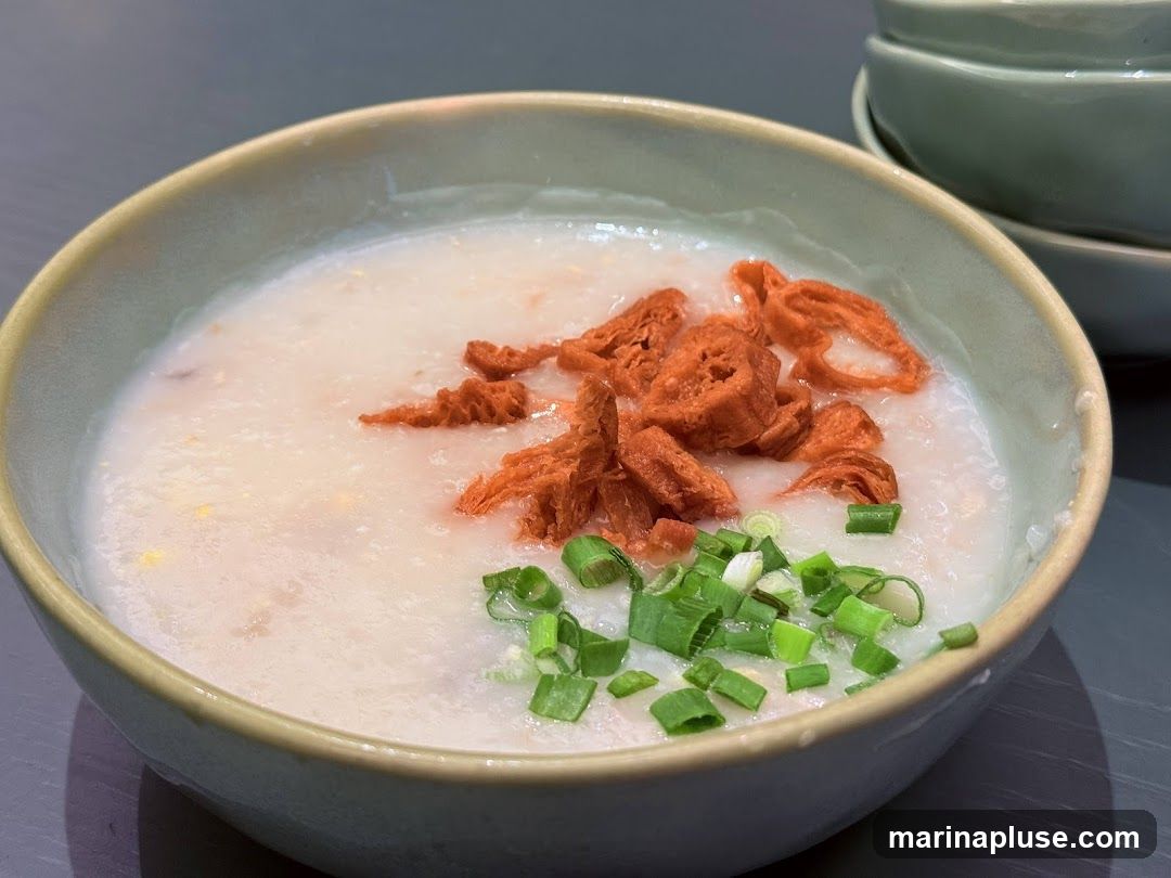 A close-up shot of a bowl of congee, a rice porridge, topped with crispy fried dough fritters and chopped scallions. The congee appears smooth and creamy, with small pieces of meat or other ingredients mixed in. The bowl is light green with a darker rim. Another stack of similar bowls is visible in the background.