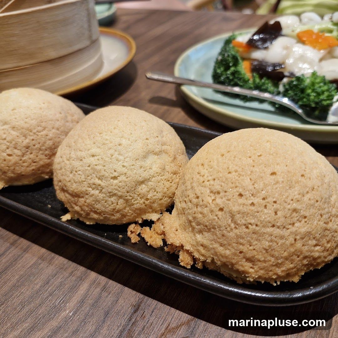 Three golden-brown baked buns with a slightly crisp exterior are presented on a black rectangular plate. In the background, a plate of stir-fried vegetables with broccoli, carrots, and mushrooms is visible, along with a bamboo steamer. The setting appears to be a restaurant with a wooden table.