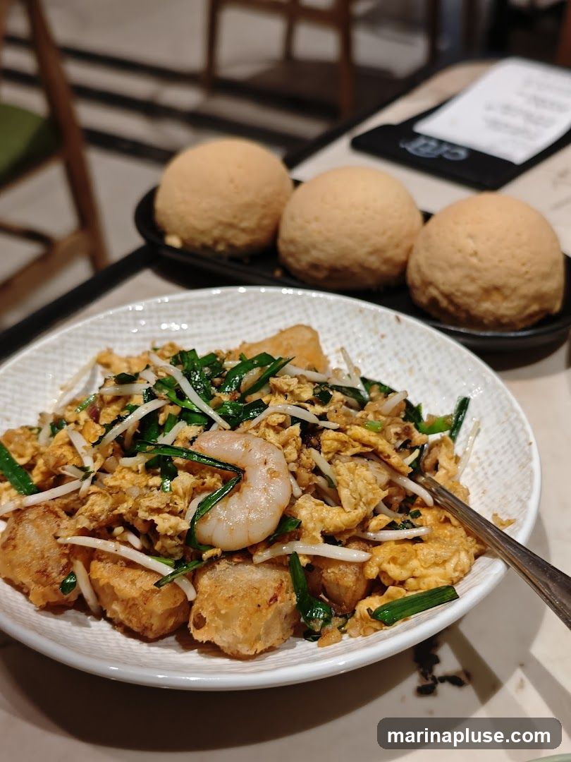 A close-up shot of a dish featuring fried turnip cake cubes, scrambled eggs, shrimp, bean sprouts, and chopped scallions. Three baked buns are visible in the background on a black plate. A menu or placemat is also visible on the table.