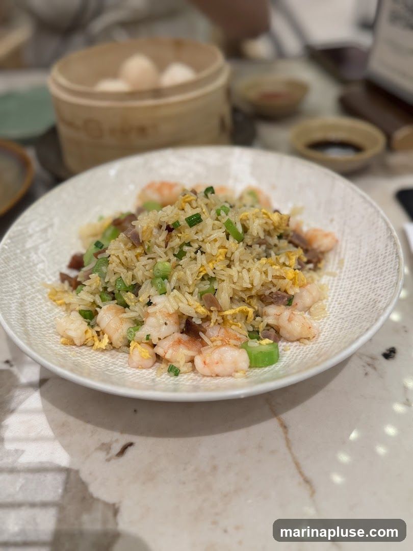 A close-up shot of a plate of shrimp fried rice, garnished with chopped green onions. In the background, a bamboo steamer basket holds several steamed buns, and small dishes of dipping sauces are visible. The food is served on a patterned plate atop a marble-patterned table.