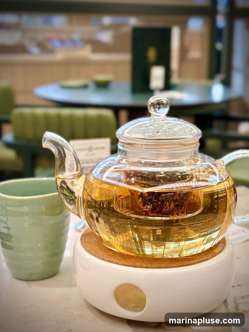 A glass teapot filled with amber tea sits on a white warmer with a cork coaster. A green teacup is to the left of the teapot. In the background, out of focus, are green chairs and tables, suggesting a cafe or restaurant setting.