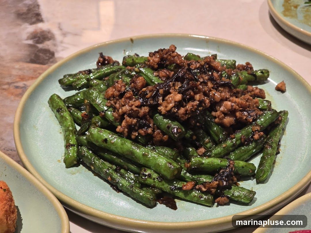 A close-up shot of a plate of sautéed green beans topped with minced meat and dark seasonings. The beans are bright green and appear slightly glossy, suggesting they have been stir-fried. The minced meat is finely crumbled and mixed with what looks like dried chilies or other spices, giving it a dark, textured appearance. The dish is served on a light green, rimmed plate, with hints of other dishes visible in the foreground and background. The table surface has a marble-like pattern.