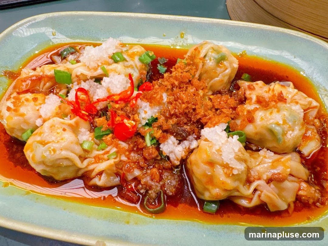 A plate of wontons in a spicy chili oil sauce, garnished with chopped scallions, crispy garlic, and chili slices.