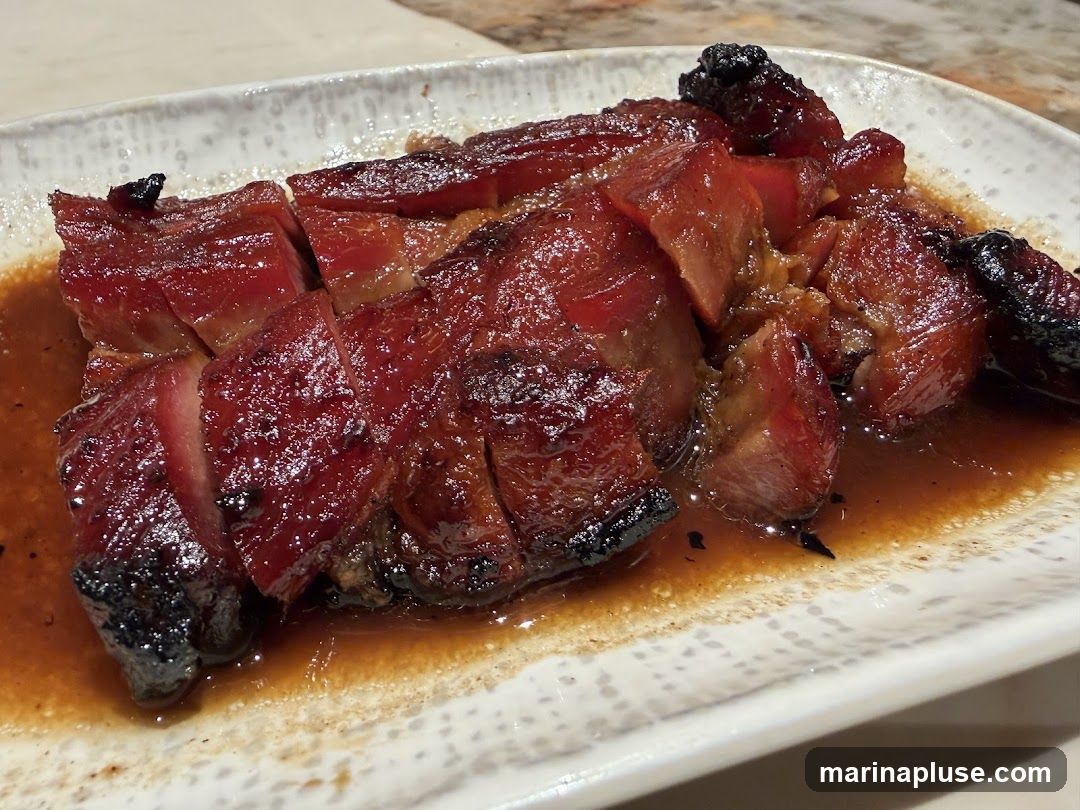 A close-up shot of sliced char siu pork, a Cantonese barbecued pork dish, served on a white patterned plate with a glossy sauce. The pork is a deep reddish-brown color with visible charring on some edges, indicating it has been roasted or grilled. The meat appears tender and juicy, with layers of fat and lean meat. The sauce is a rich brown, likely a glaze made from soy sauce, honey, and spices.