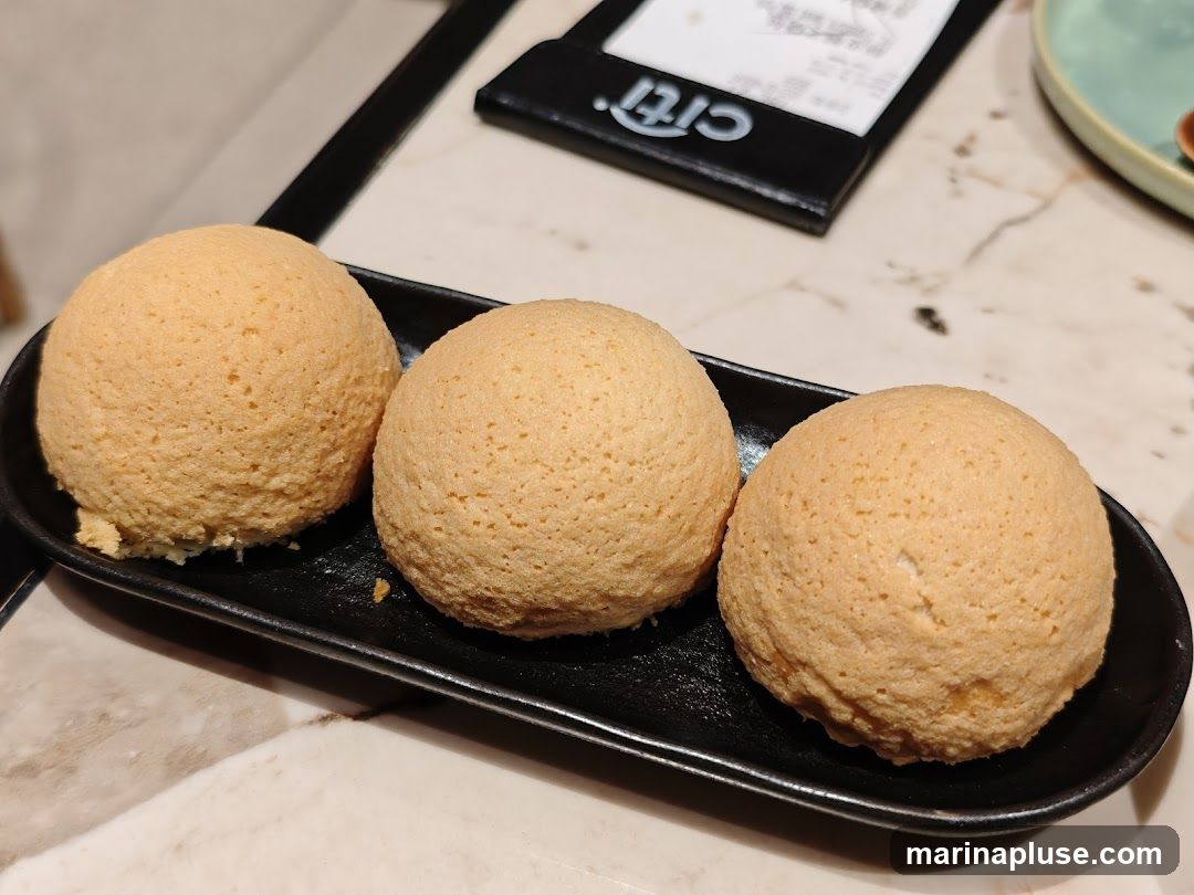 Three golden-brown baked buns are served on a black oval plate. The buns have a slightly textured surface and appear to be soft. In the background, a menu with the word "Citi" is visible, along with a portion of a light green plate. The table surface has a marble-like pattern.