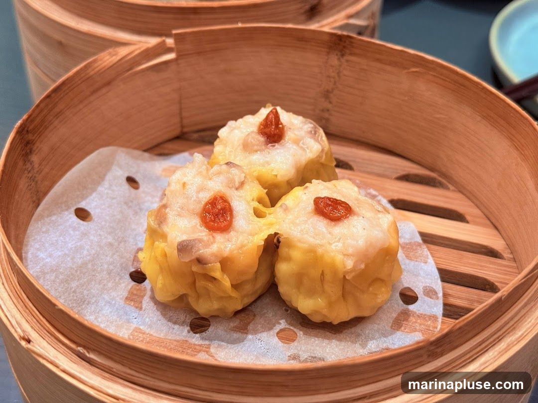 Three steamed dumplings, likely siu mai, are presented in a bamboo steamer lined with parchment paper. The dumplings have a yellow wrapper and are topped with a small red garnish.