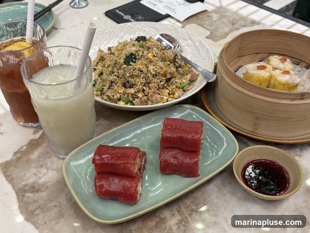 A table setting with various dim sum dishes and drinks. In the foreground, there are red rice noodle rolls served on a light green plate, with a small bowl of dark sauce. Behind the rolls, a plate of fried rice is visible, and to the right, a bamboo steamer contains several pieces of siu mai. Two glasses with drinks, one appearing to be iced tea with lemon and the other a milky beverage, are on the left side of the image. A menu or bill is partially visible in the background.