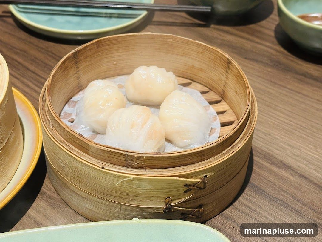 A close-up shot of a bamboo steamer filled with four delicate, translucent steamed dumplings. The dumplings have a pleated appearance and appear to be filled with shrimp or a similar filling. The steamer is placed on a wooden table, with other dishes and chopsticks visible in the background.