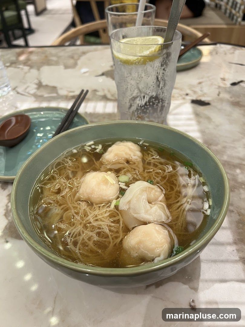 A bowl of wonton noodle soup with several wontons and thin noodles in a clear broth, garnished with chopped green onions. A glass of sparkling water with lemon slices and ice is in the background, along with chopsticks and a small dish. The table has a marble-like pattern.