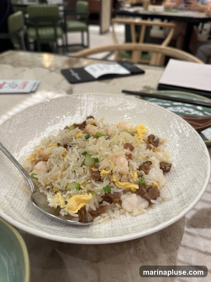 A close-up shot of a bowl of shrimp fried rice, with a spoon resting on the side of the bowl. The rice is mixed with shrimp, scrambled eggs, and green onions. In the background, a blurred view of a restaurant interior with tables and chairs is visible.