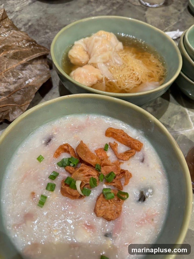 A close-up shot of two bowls of food. In the foreground is a bowl of congee topped with fried shallots and chopped scallions. In the background is a bowl of wonton noodle soup with several wontons and thin noodles in broth. A large dried leaf is visible to the left of the bowls.