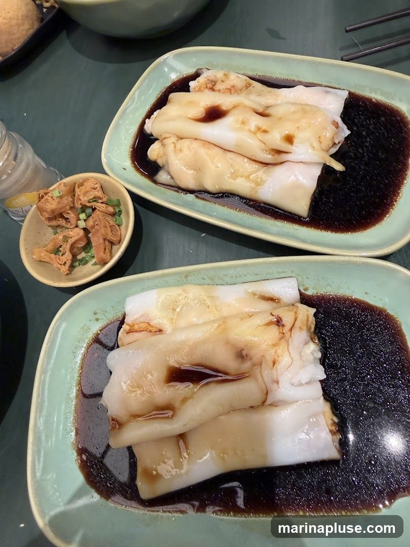 Two plates of steamed rice rolls, also known as cheung fun, are served with a dark soy sauce. A small dish of fried dough fritters, possibly youtiao, garnished with chopped green onions is also visible. The setting appears to be a restaurant table with a dark green surface.