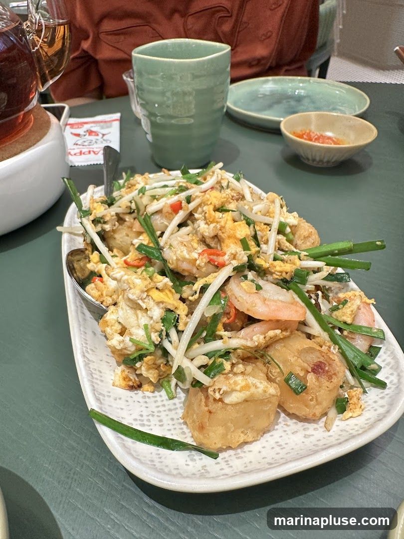 A plate of Pad Thai with shrimp, scrambled eggs, bean sprouts, and green onions is served on a white textured plate. A small dish of dipping sauce, a green ceramic cup, and a glass of amber liquid are also visible on the table. A small packet with a red logo is to the left of the cup.