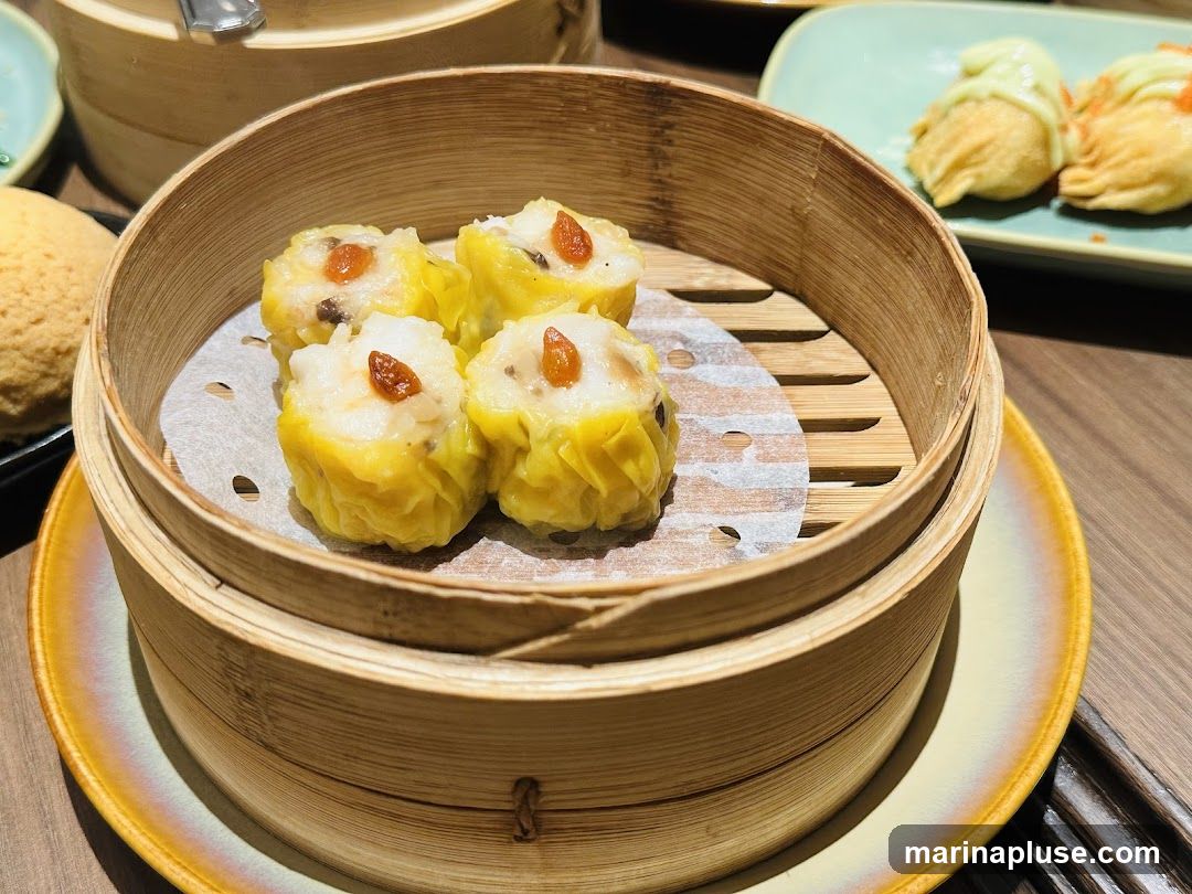 A close-up shot of four steamed dumplings in a bamboo steamer basket. The dumplings are yellow and topped with a small piece of red garnish. In the background, another bamboo steamer and a plate with two fried dumplings are visible.