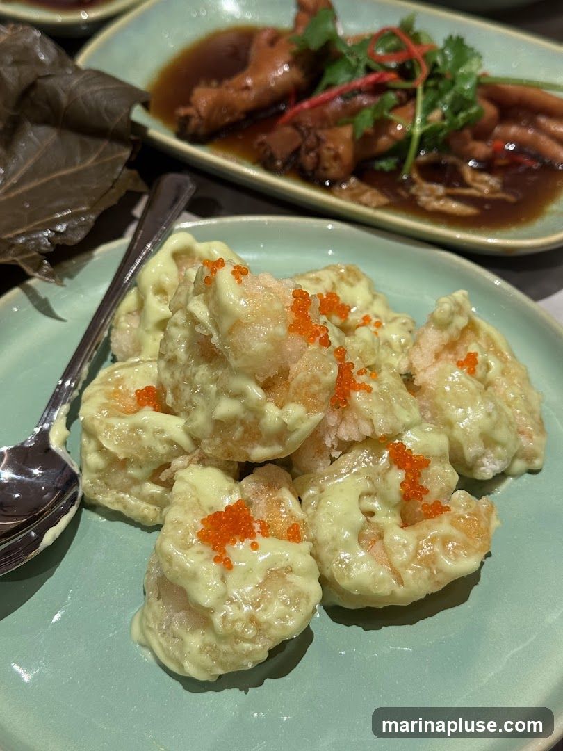 A close-up shot of a plate of mayonnaise shrimp, garnished with bright orange roe. The shrimp are coated in a creamy, pale green sauce and appear to be lightly fried. In the background, another dish of braised chicken feet is visible, served in a dark sauce with cilantro and chili threads. A large, dried lotus leaf is also present on the left side of the frame, adding a rustic touch. A spoon rests on the plate of shrimp.