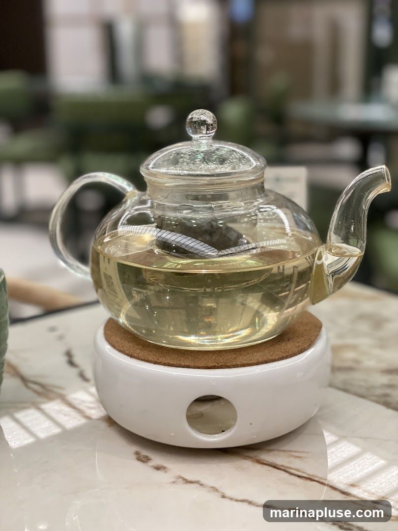 A clear glass teapot filled with light-colored tea sits on a white ceramic tea warmer with a cork coaster. The teapot has a glass lid with a decorative knob and a glass handle and spout. An infuser basket is visible inside the teapot, holding tea leaves. The tea warmer has a circular opening on the front. The teapot and warmer are placed on a marble-patterned surface. The background is blurred, showing hints of green seating and other tables, suggesting an indoor cafe or restaurant setting.