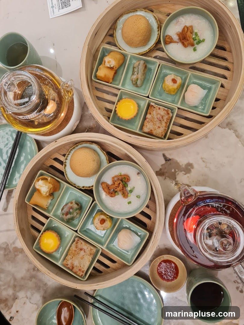 A top-down view of a table laden with a dim sum meal. Two bamboo steamers are filled with various small dishes, including spring rolls, dumplings, tarts, and congee. Teapots filled with amber liquid and cups are also visible, along with small dishes of dipping sauce. The meal is presented on a marble-patterned tabletop.