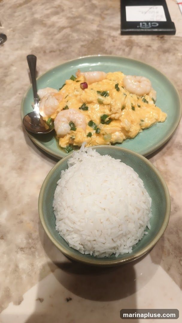 A plate of scrambled eggs with shrimp and chopped green onions is served with a bowl of white rice. A spoon rests on the plate. A small black object with text is visible in the background.