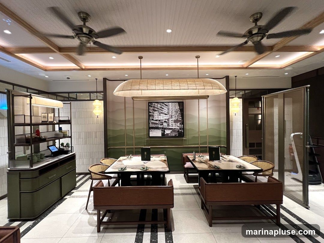 A well-lit restaurant interior with two tables set for dining. The tables have chairs and banquette seating. A large black and white framed picture hangs on the wall behind the tables, which features a cityscape. There are ceiling fans and pendant lights. To the left, a counter area with shelves displays dishes and a laptop. The overall decor appears modern with some traditional elements.