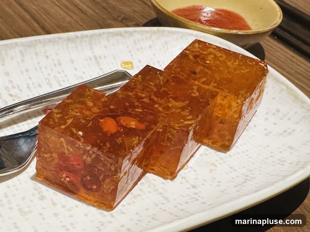 Three cubes of amber-colored jelly dessert are arranged on a white, textured plate. The jelly appears to contain small pieces of fruit or herbs, including visible goji berries and possibly red chili slices. A small bowl with a red sauce is visible in the background, along with cutlery.