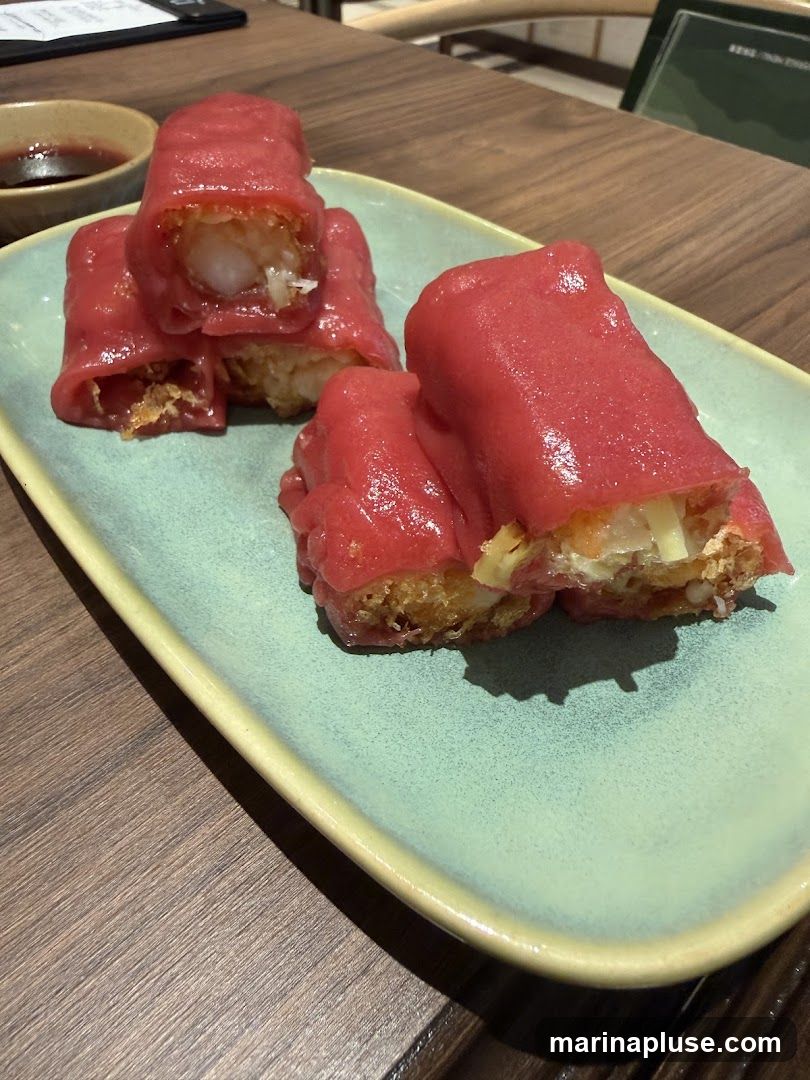 A plate of pink shrimp rice rolls with a crispy fried filling, served on a light green rectangular plate. A small bowl of dark dipping sauce is visible in the upper left corner. The rolls are cut in half, revealing the shrimp and other ingredients inside. The dish is placed on a wooden table.