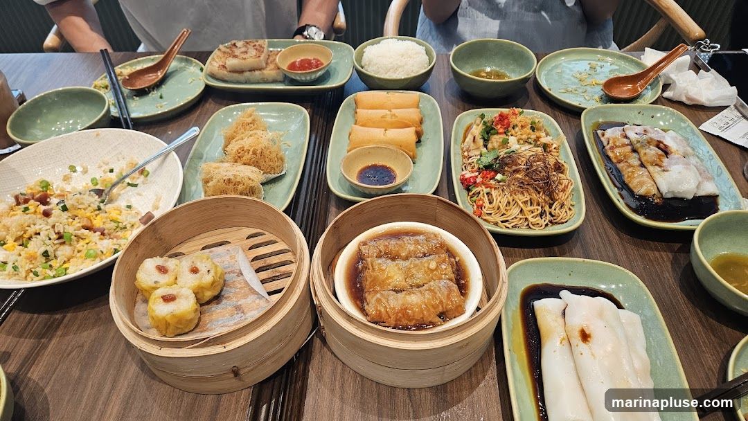 A table laden with various dim sum dishes, including fried rice, steamed dumplings (siu mai), spring rolls, noodles, and rice noodle rolls, all served in traditional bamboo steamers and ceramic plates.