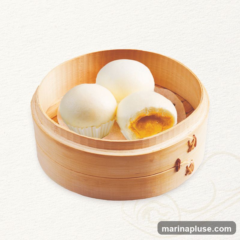 Three white steamed buns are presented in a bamboo steamer. One bun is cut in half, revealing a bright yellow filling.